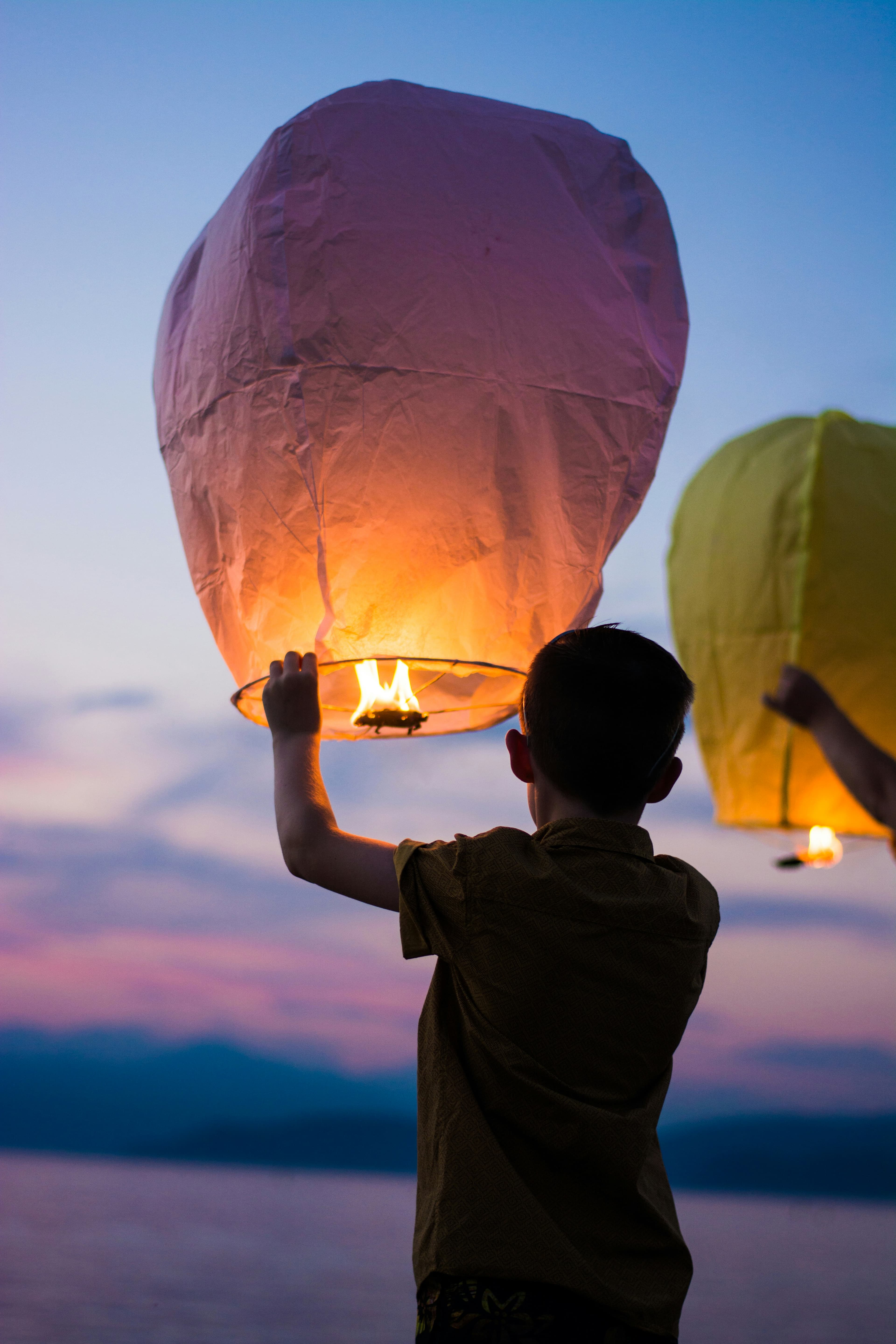 Deux enfants se tiennent devant un coucher de soleil, tenant des lanternes célestes aux couvertures jaune et rose. La personne à gauche regarde vers les lanternes, tandis que celle à droite regarde vers l'appareil photo. Ils sont près d'un plan d'eau lors d'une activité de soirée festive.