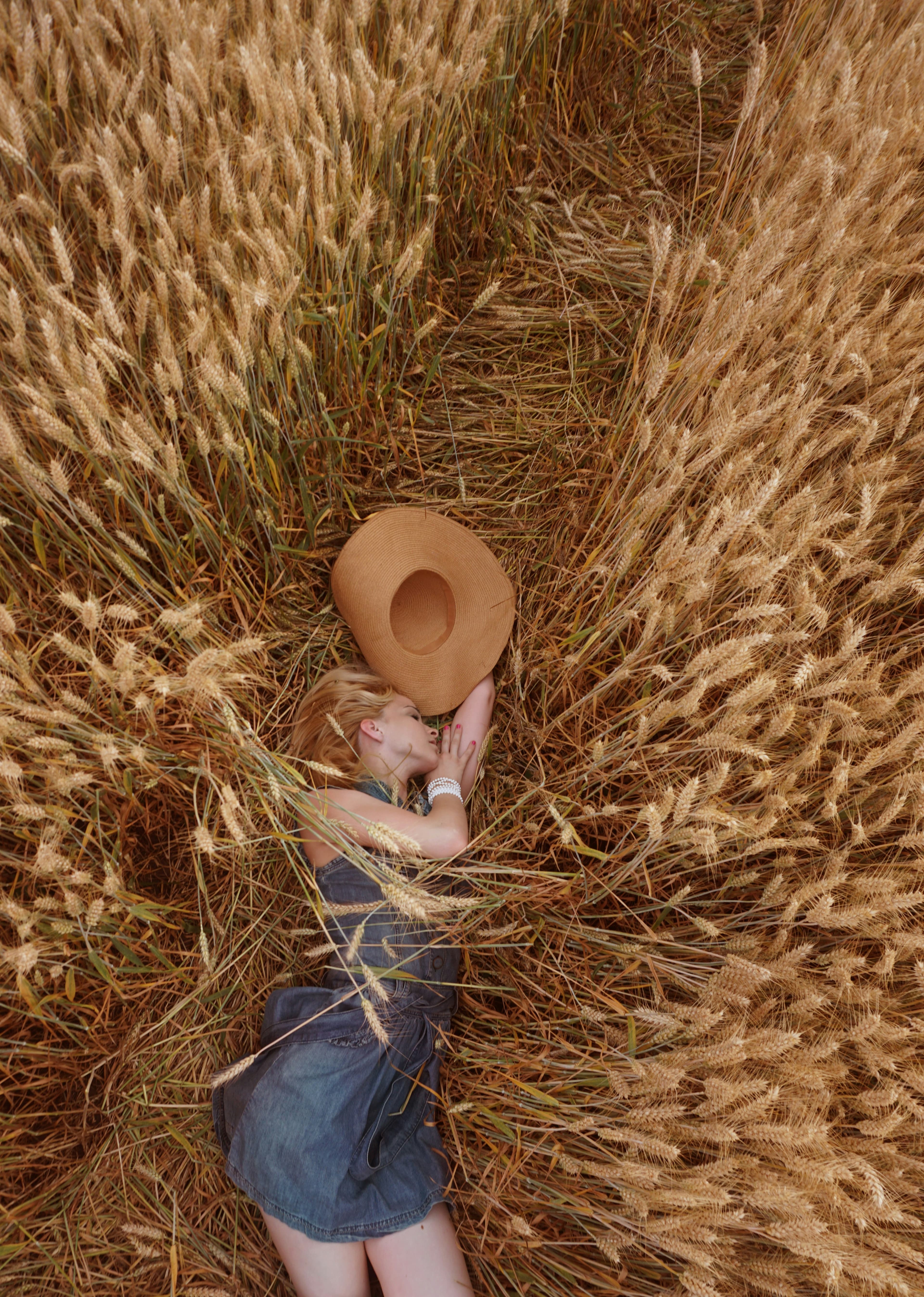 Une femme allongée paisiblement dans un champ de blé doré. Elle porte un chapeau clair en paille et un haut en denim foncé. Son bras est tendu vers l'extérieur, comme pour saisir quelque chose. Des épis de blé au premier plan masquent partiellement la personne, tandis qu'elle est allongée sur des épis de blé allongés.