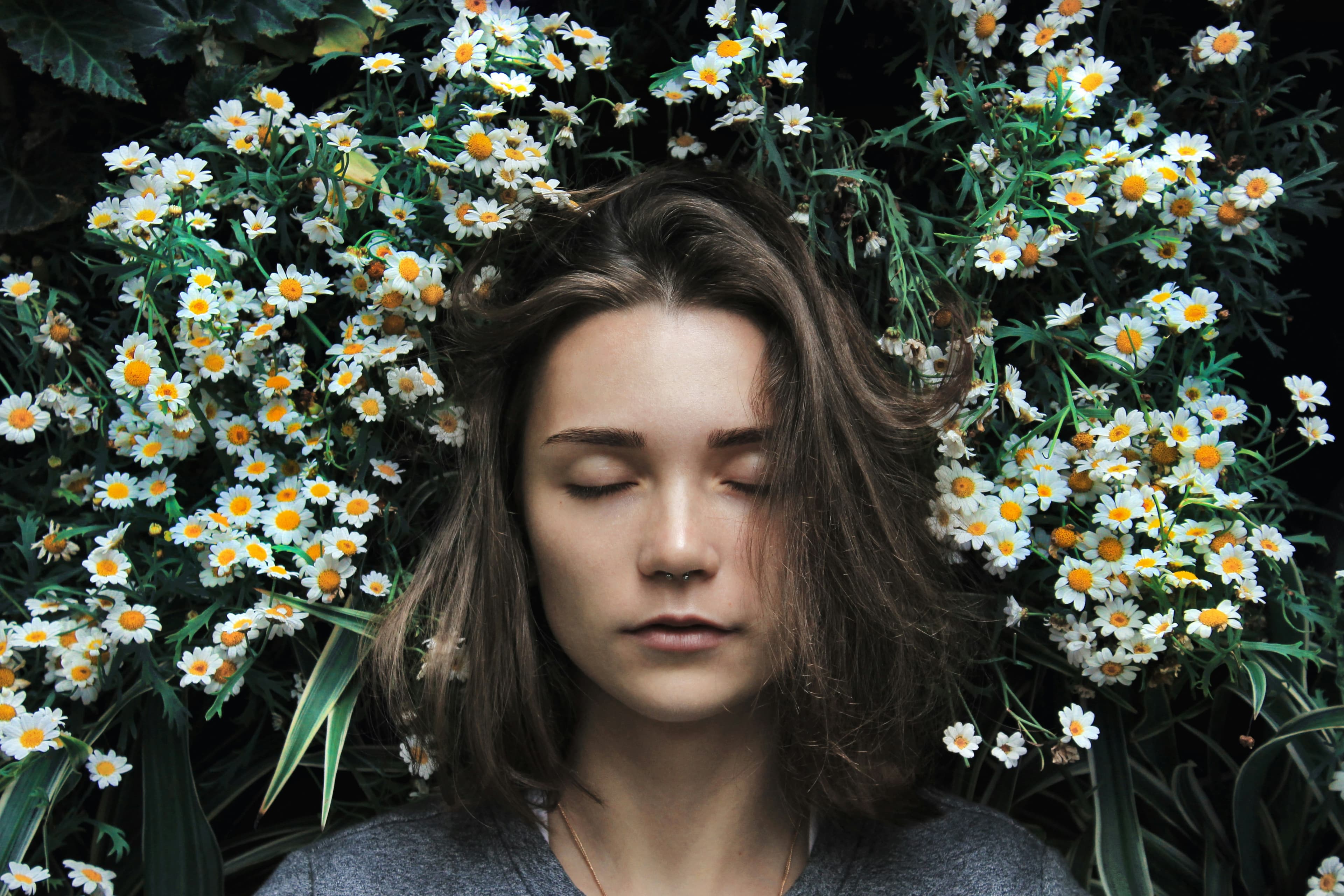 Portrait artistique d'une jeune femme au teint clair et aux cheveux bruns, les yeux fermés dans une expression sereine. Son visage est entouré de fleurs de marguerites éclatantes sur un fond verdoyant, créant une atmosphère onirique et harmonieuse. 